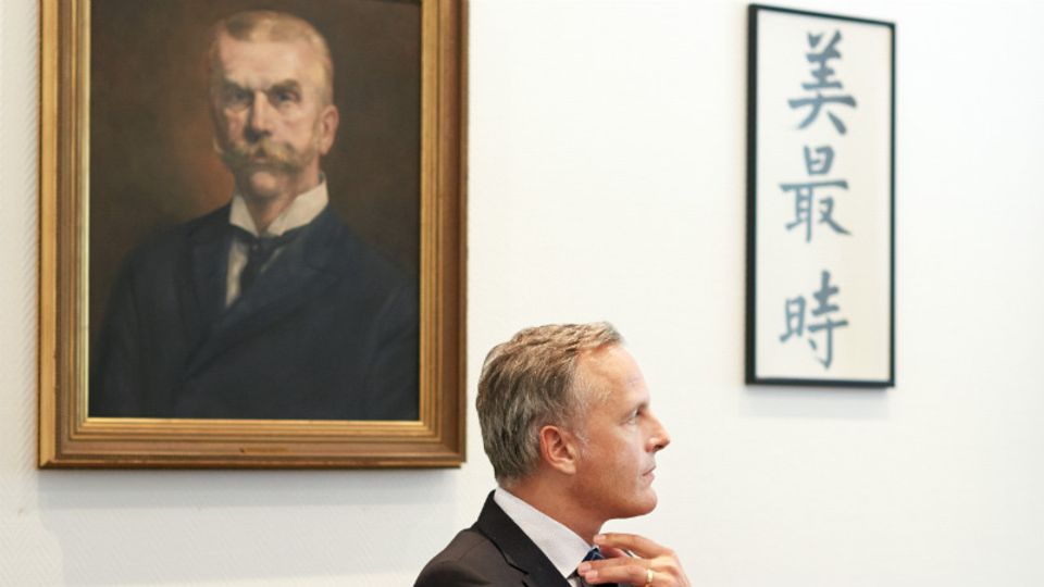 Dirk Sänger unter dem Porträt von Hermann Melchers (1842–1918), der 1866 das Geschäft in Hongkong aufbaute. Daneben steht der Firmenname in chinesischen Schriftzeichen. Übersetzt bedeuten sie „ewige Schönheit“