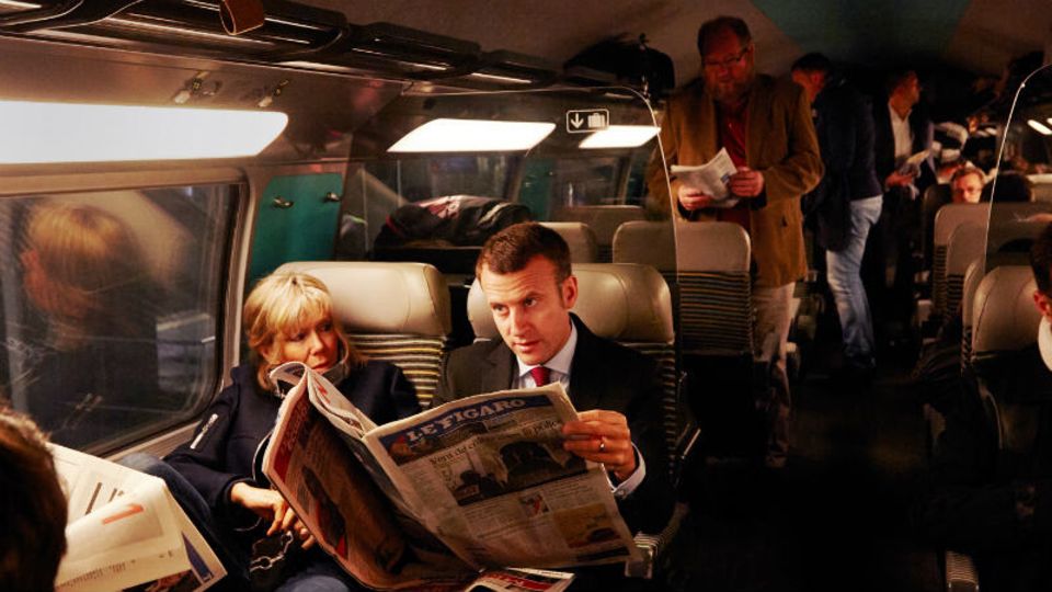 Macron und seine Frau Brigitte lesen, was die Zeitungen über seinen Auftritt am Vortag schreiben. Seit er nicht mehr Minister ist, reist er öfter zweiter Klasse. Da er keine Partei im Rücken hat, muss gespart werden