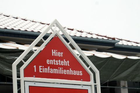 Ein Schild mit der Aufschrift "Hier entsteht ein Einfamilienhaus" steht vor einem eingerüsteten Rohbau