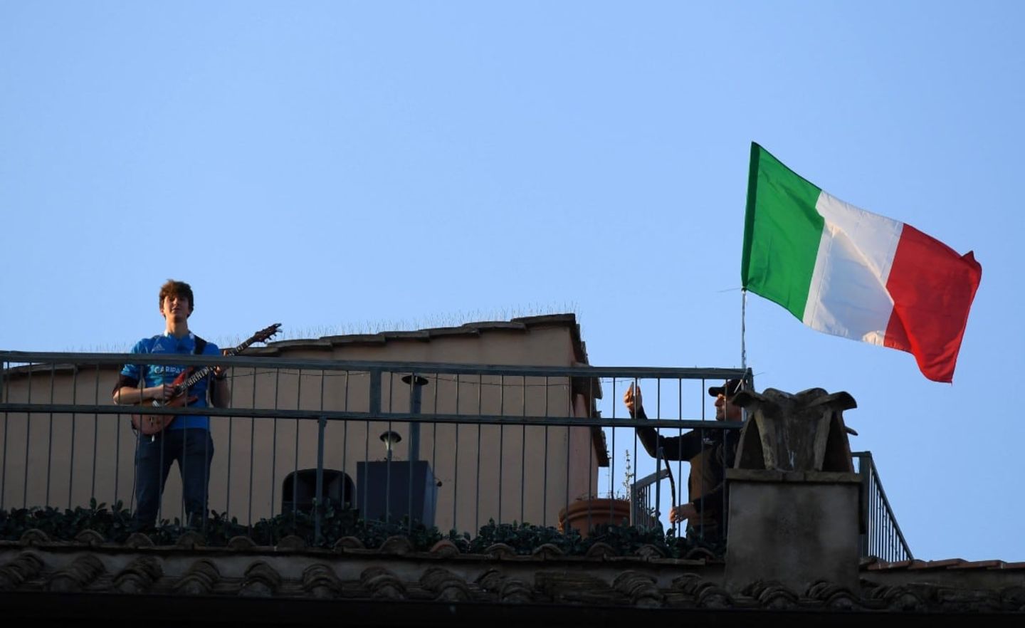 Ein Mann steht auf einem Dach und spielt Guitarre, neben ihm weht die italienische Flagge