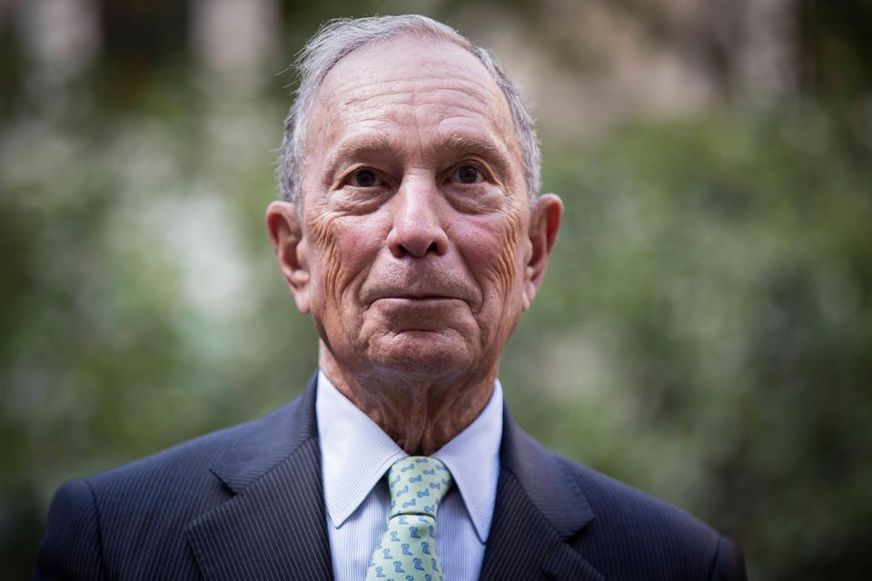 Michael Bloomberg vor einem unscharfen, grünen Hintergrund mit schwarzem Jackett, blauem Hemd und Krawatte
