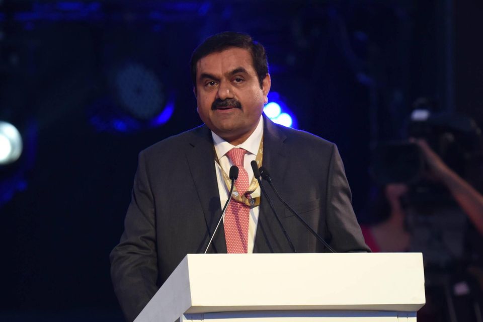 Gautam Adani steht hinter einem Pult mit Mikrofonen. Er trägt ein schwarzes Jackett, ein weißes Hemd und eine rote Krawatte.