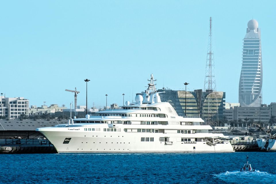 Die weiße Yacht liegt in einem Hafen mit der Skyline der Stadt im Hintergrund – ein Turm zeichnet sich am Horizont ab