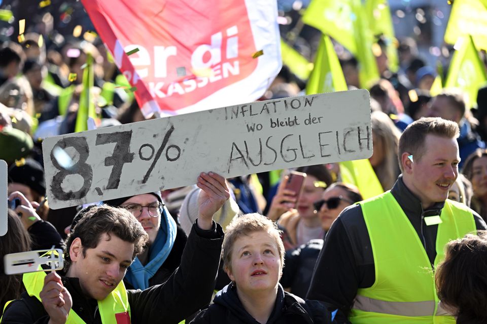 Streikende stehen mit einem Schild mit der Aufschrift "87% Inflation - Wo bleibt der Ausgleich"  auf einem Platz