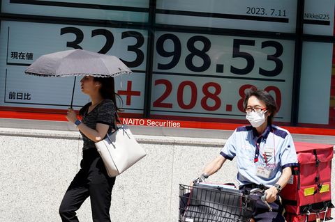 Eine Frau geht mit einem aufgespannten Schirm an einer elektronischen Anzeigentafel mit Kursdaten des Nikkei-Index vorbei