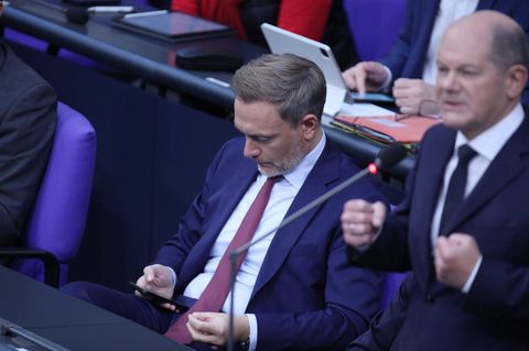 Bundesfinanzminister Christian Lindner liest in seinem Smartphone, rechts steht Bundeskanzler Olaf Scholz am Mikrofon