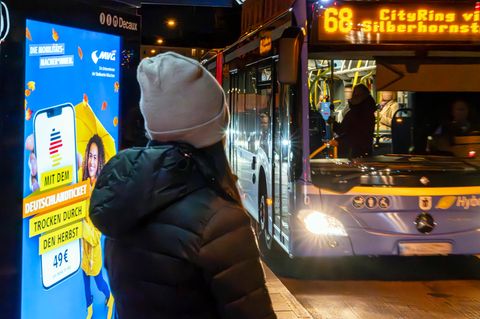 Frau wartet auf den Bus, neben ihr Leuchtwerbung für das Deutschlandticket
