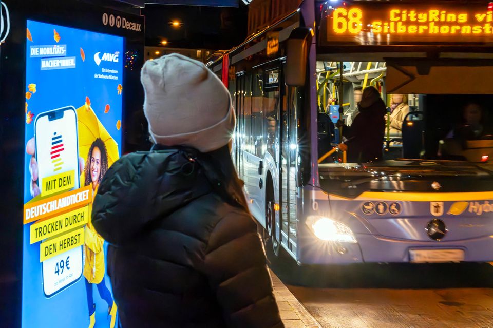 Frau wartet auf den Bus, neben ihr Leuchtwerbung für das Deutschlandticket