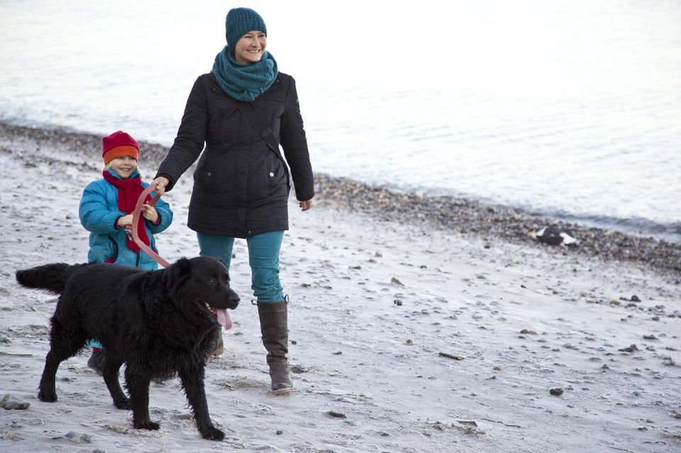 Eine Frau geht mit einem Kind und einem schwarzen Hund am Strand spazieren