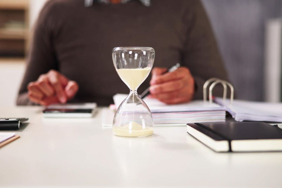 Eine Sanduhr steht auf einem Schreibtisch, an dem jemand arbeitet