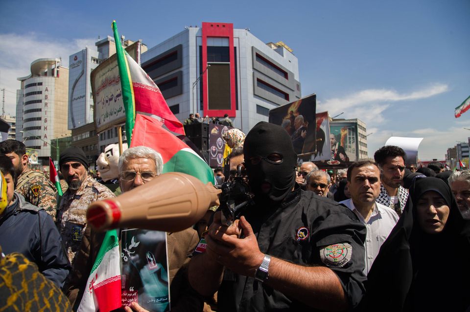 Soldat der Revolutionsgarde im Iran mit Sturmmaske steht in Menschenmenge und zielt mit seiner Panzerfaust auf die Kamera