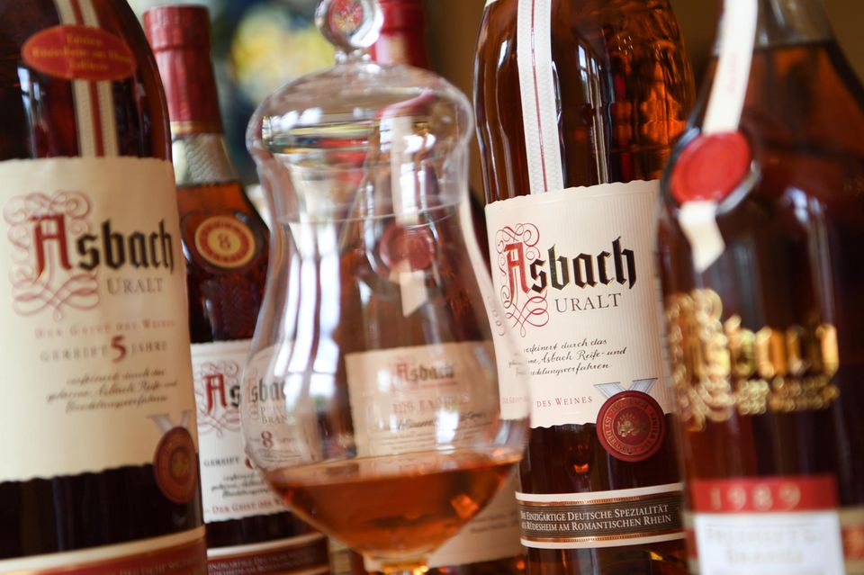 A glass of brandy is placed in front of bottles of various Asbach-Uralt brandies