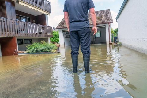Ein Mann steht mit Gummistiefeln vor einem Haus und einer Garage, die unter Wasser stehen