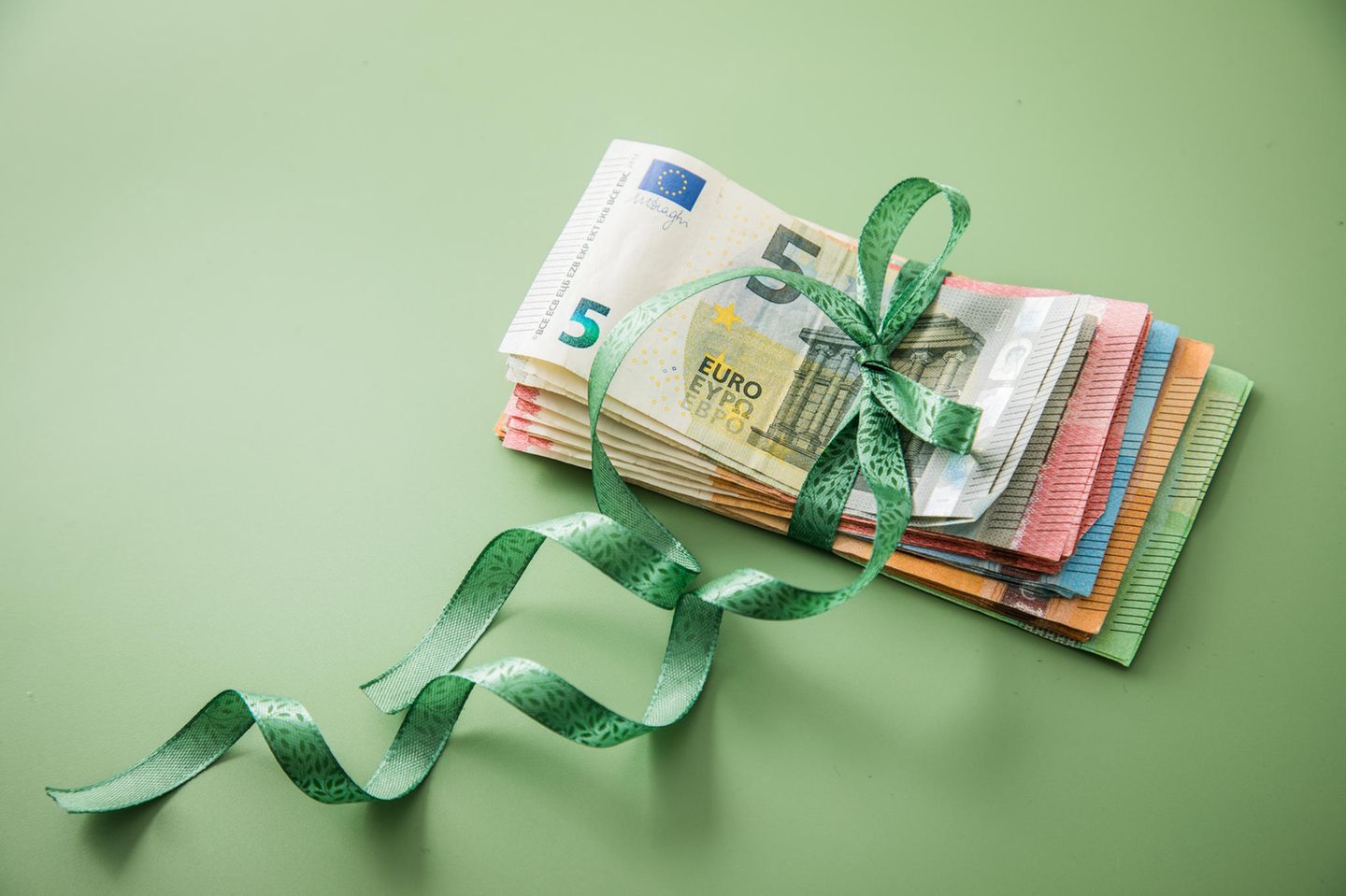 Ein zusammengelegter Geldstapel aus unterschiedlichen Scheinen ist mit einer grünen Schleife zusammengebunden