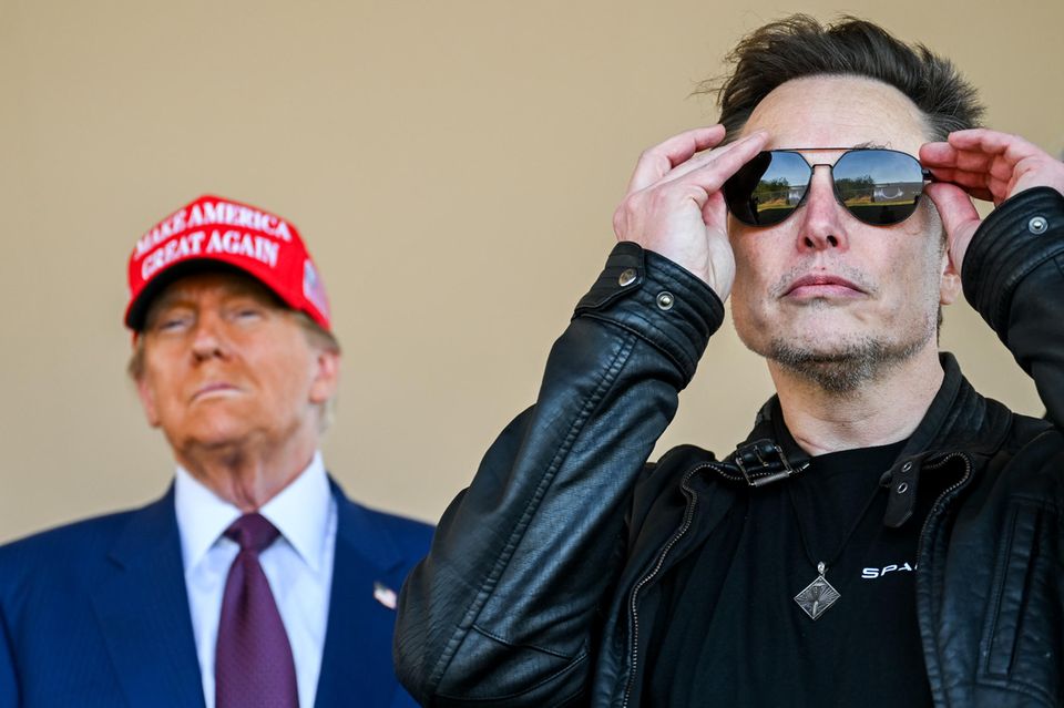 Elon Musk setzt sich eine Sonnenbrille auf, während Donald Trump im Hintergrund steht