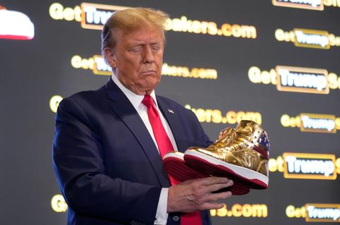 Donald Trump hält gold glänzende Sneaker in der Hand und betrachtet diese.