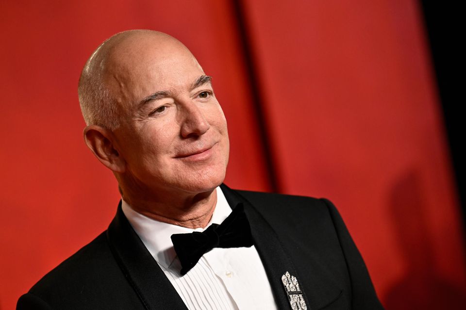 Jeff Bezos steht im Smoking for einer roten Wand.