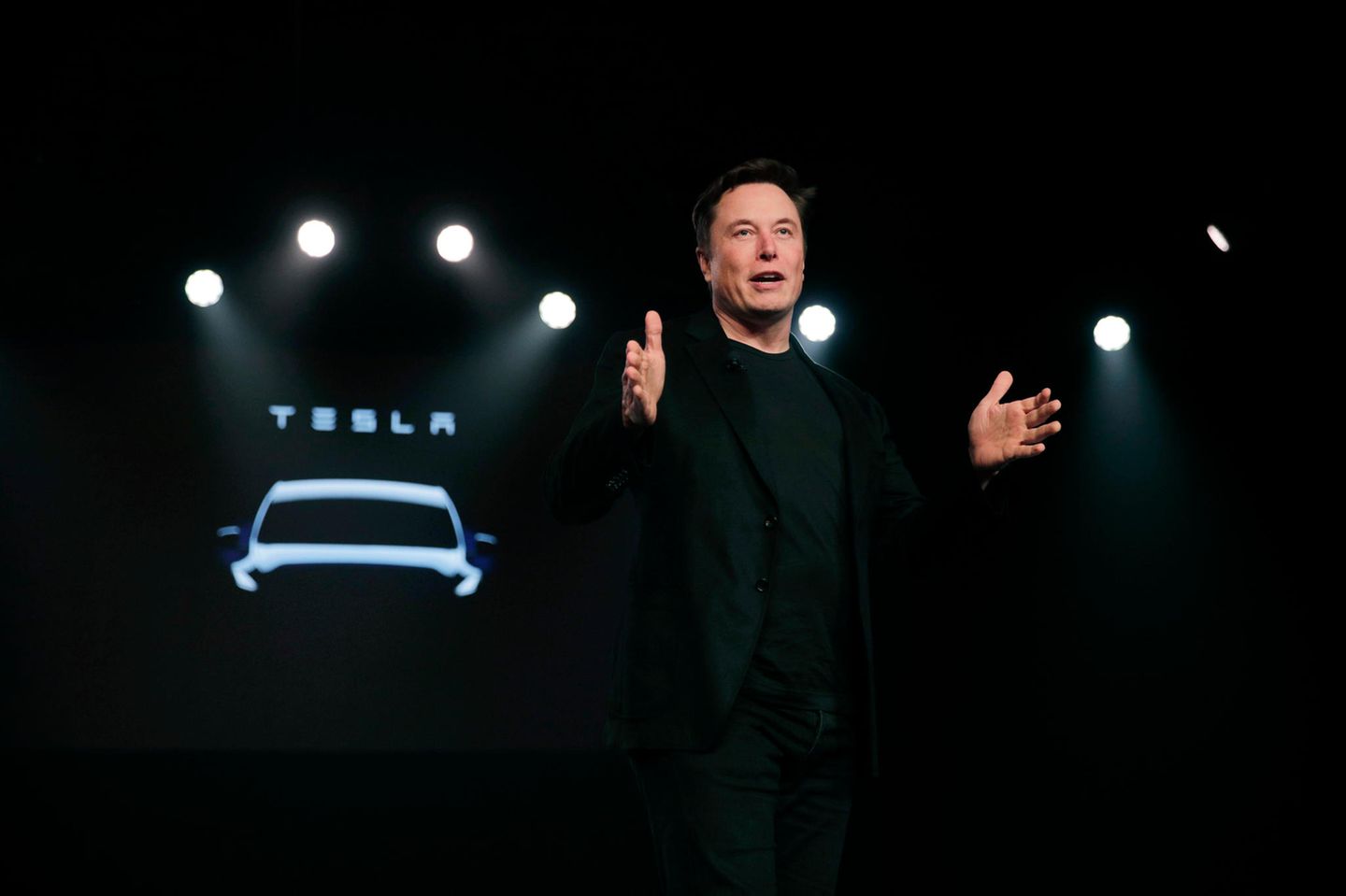 Elon Musk präsentiert auf einer dunklen Bühne ein neues Tesla-Auto, das im Hintergrund bereits als Silhouette angedeutet ist