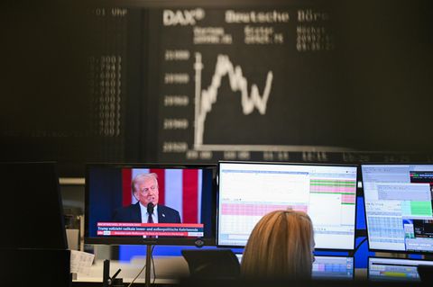 Dax-Kurve zeigt im Handelssaal der Frankfurter Börse, während auf einem Bildschirm Trump zu sehen ist
