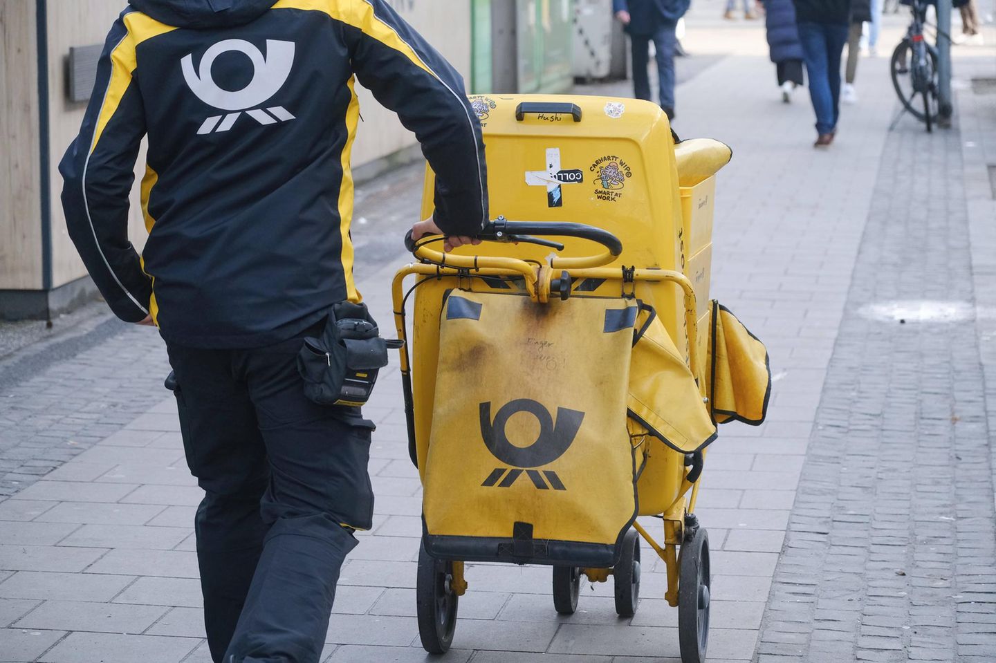 Ein Briefzusteller schiebt seinen Trolley über einen Gehweg