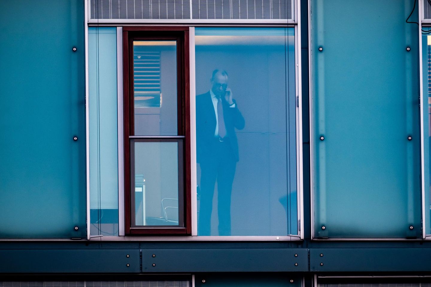 Friedrich Merz steht telefonierend hinter einer Glasscheibe