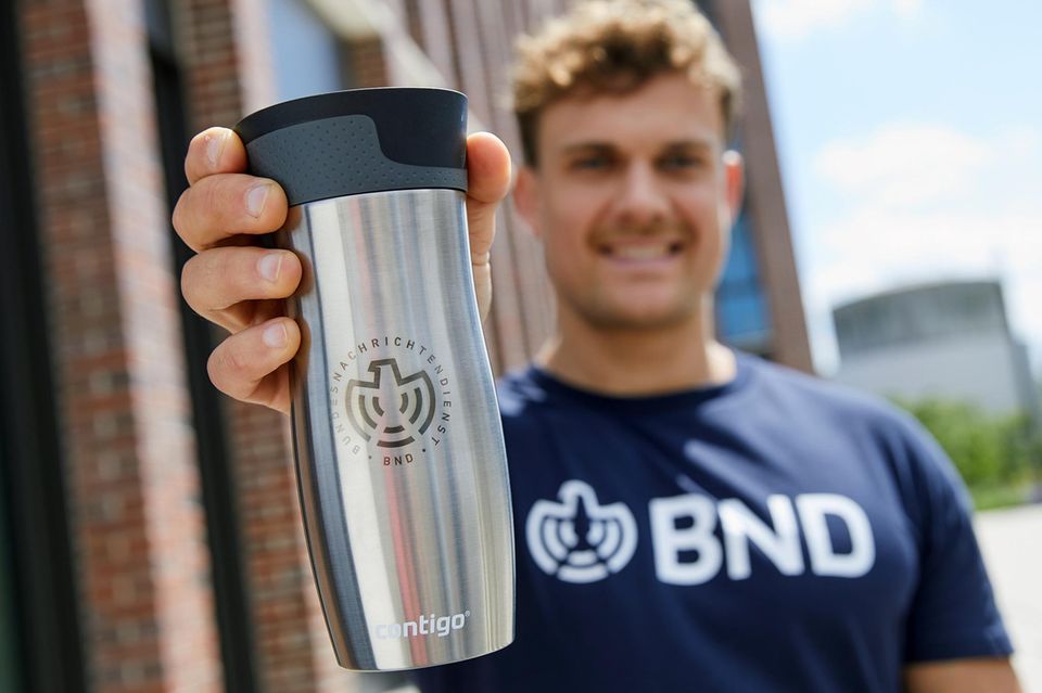 In einem Werbefoto hält jemand einen silbernen Kaffeebecher mit BND-Logo in die Kamera