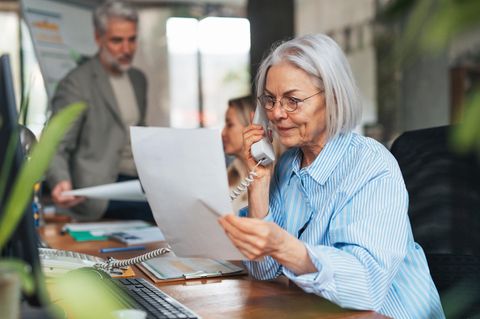 Eine ältere Frau sitzt im Büro und telefoniert mit einem Blatt Papier in der Hand