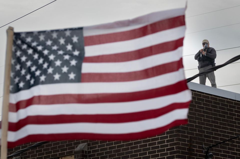 Ein ICE-Beamter steht auf einem Hausdach vor dem eine US-Flagge weht