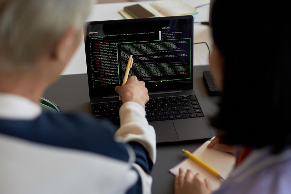 Ein älterer Herr zeigt auf einen Laptop, auf dem Codereihen laufen